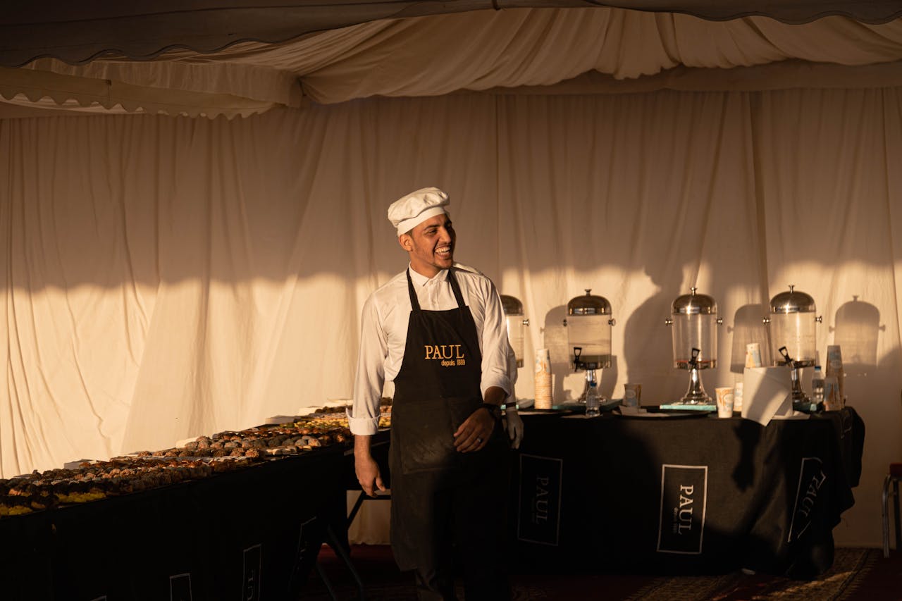 A smiling chef in an apron stands at a buffet table with pastries, creating a welcoming atmosphere.